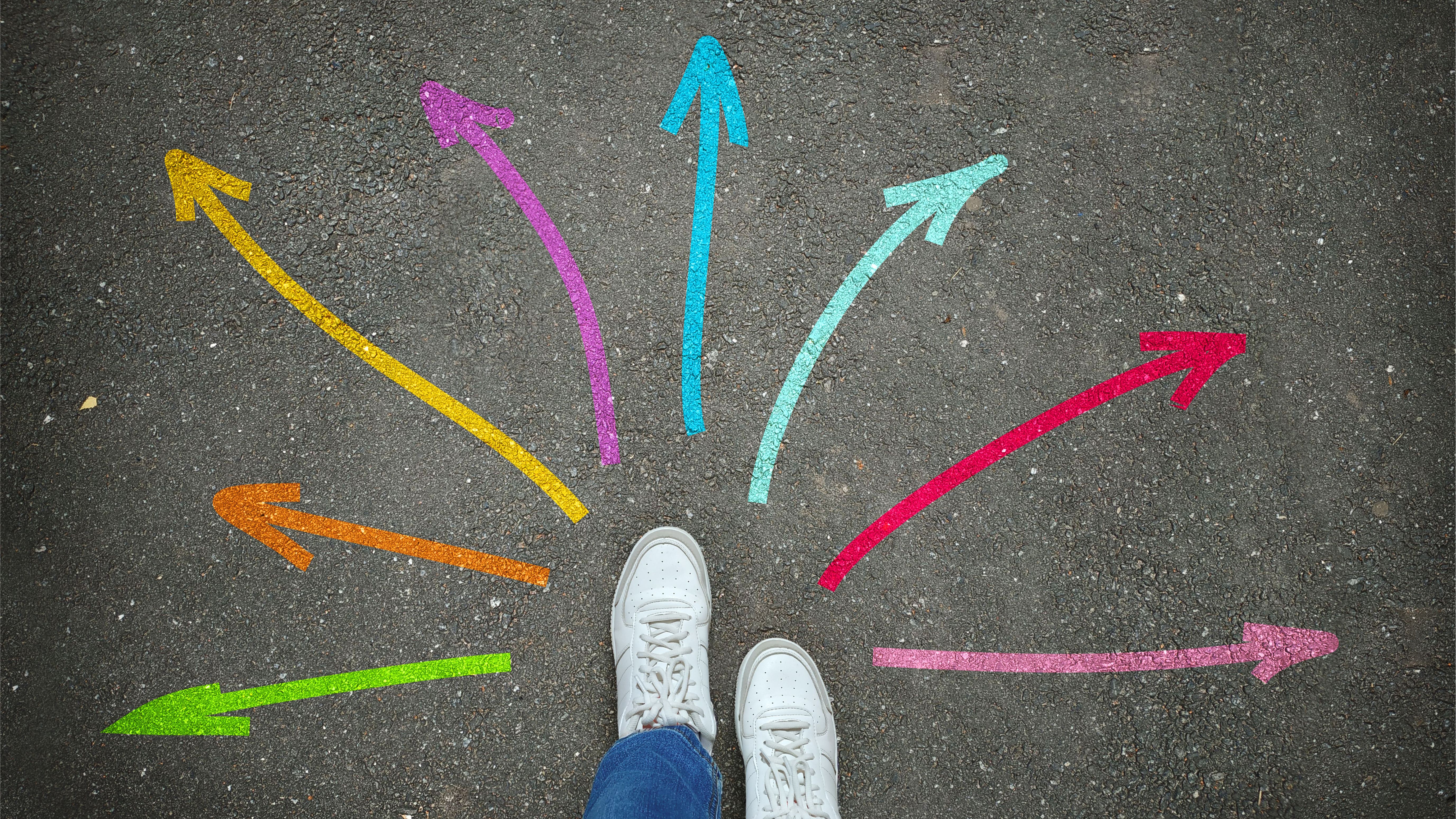 Benen på vita tennisskor ligger i grått framför färgade pilar som pekar i olika riktningar på asfalten
